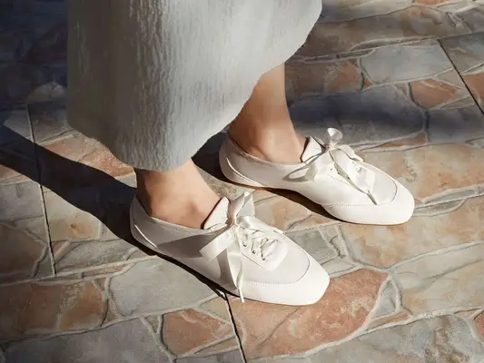 Feet wearing elegant white sneakers with ribbon ties, standing on a tiled floor