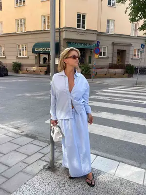 Matilda Djerf wearing a light blue button-down shirt with matching cotton maxi skirt, sunglasses, white handbag, and flip flops