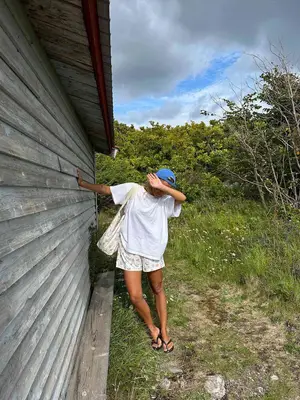 Matilda Djerf wears a blue baseball cap, oversized white t-shirt, white shorts, a white bag, and black flip-flops