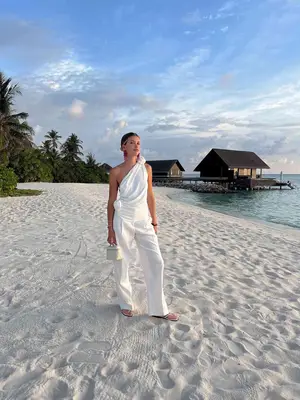 Sofia Richie Grainge wears white top with a flower shoulder detail, white pants, and silver flip-flops on beach