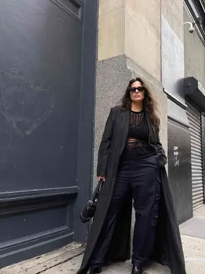 Ashley Graham wears black sunglasses, long black trench coat, black pants, black shirt, and black purse