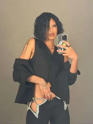 Willow smith taking mirror selfie wearing black buttoned vest, jacket on arms, pants with silver rhinestone cutouts, rings, and smoky eye makeup