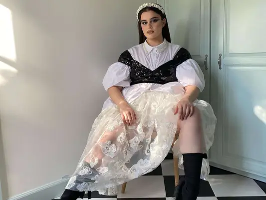 Barbie Ferreira wears a white button-down puff-sleeve top, black sequin crop top, lacy white skirt, black shoes and socks, and white pearl headband