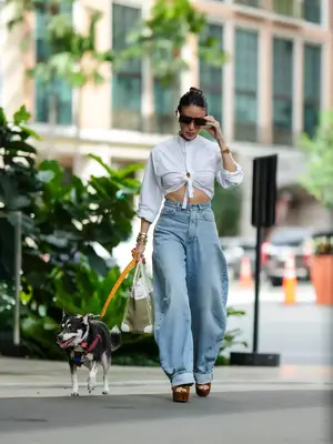 Woman wearing white tied cropped blouse, horseshoe light wash jeans, platform shoes, sunglasses, and walking dog