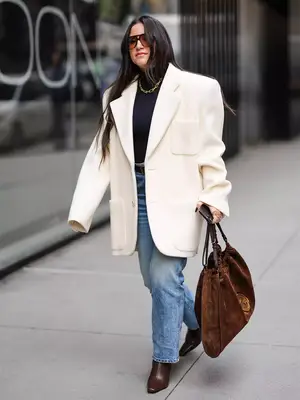 Woman wears black turtleneck, light wash jeans, oversized white blazer, brown leather handbag, chain necklace, and aviator sunglasses