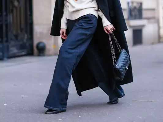 Woman wearing flared wide leg dark wash jeans, white sweater, long black coat, puffy Chanel bag, and black shoes