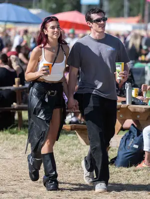 Dua Lipa wears black-and-white outfit of leather jacket, boots, white tank top, and silver jewelry while attending Glastonbury festival with Callum Turner