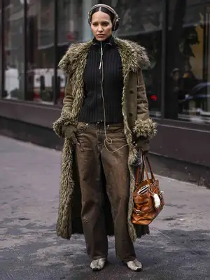 Model wears brown jeans, green faux fur coat, black sweater, tan belted leather handbag, and over-ear headphones