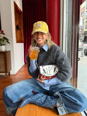 Content creator Izzi wears cuffed jeans, gray mohair sweater, denim boots, blue button-down, colorful Louboutin bag, and yellow Yankees cap