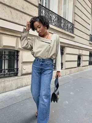 Ellie Delphine wears an asymmetrical button-down, flared jeans, open-toe shoes, and black handbag with structural handle