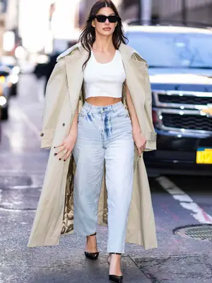 Camila Morrone wearing black sunglasses, khaki trench coat, white cropped tank, blue barrel jeans
