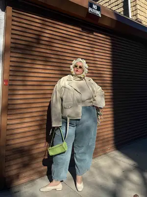 Abby Bible wears light wash wide leg jeans, a cream-colored leather-jacket, sweater, and flats, graan handbag and sunglasses, and floral hood