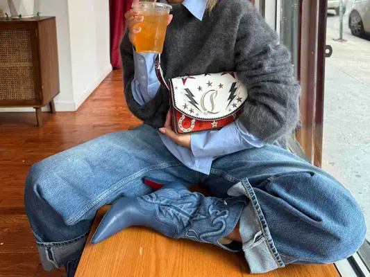Content creator Izzi wears cuffed jeans, denim boots, a light blue button-down, gray mohair sweater, and Louboutin handbag