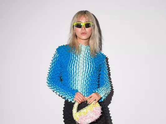 Betty Bachz wearing long-sleeve blue popcorn top, racer sunglasses, and a black skirt