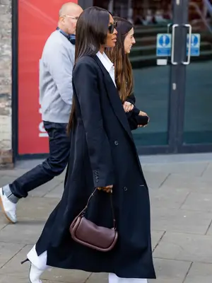 Naomi Campbell wears a black coat, white pants and button-down, and brown leather handbag