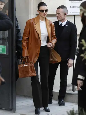 Kendall Jenner wears a brown leather blazer, white top, black trousers and boots, brown Hermes bag, and black sunglasses