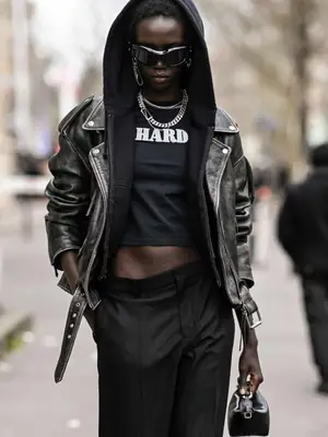 Anok Yai wearing black graphic tee that says hard layered with black sweatshirt wearing hood, black leather jacket, black sunglasses, and layered silver jewelry