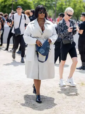 Precious Lee wears a gray trench coat, blue Dior saddle bag, and black loafers with sunglasses and chain necklace