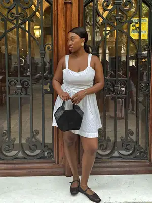 Nnenna Echem wearing a short white linen dress, black Mary Jane shoes, and a black handbag.
