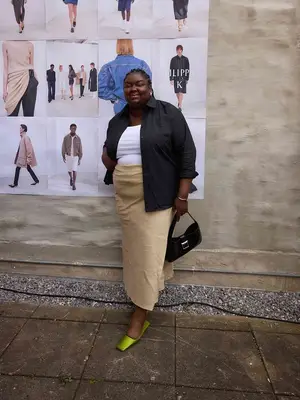 Abisola Omole wearing a black linen shirt under a white t-shirt, a tan linen maxi skirt, green slides, and holding a black purse.