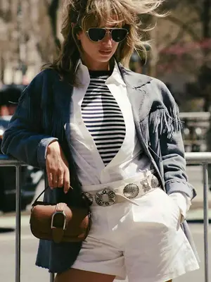 Content creator Izzi wearing a pair of sunglasses, a blue fringe jacket, a white linen shirt, a black and white stripe shirt, white linen shorts, and a white belt with silver buckles