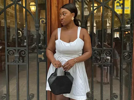 Nnenna Echem wearing a white linen mini dress, gold earrings, and black handbag