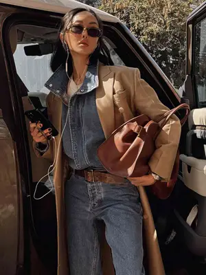 Chriselle Lim wears a gray denim jumpsuit, beige trench coat, cognac leather handbag, sunglasses, and wired headphones