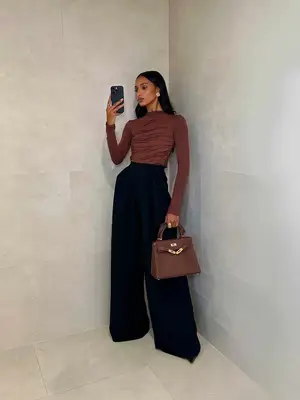 Jasmine Tookes wears a brown ruched top, black wide leg trousers, and brown handbag with gold hardware