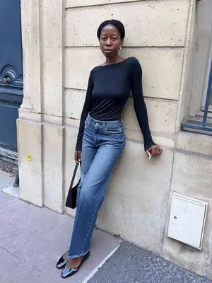 Sylvie Mus wears a black knit top, medium wash straight leg jeans, black flats, and black shoulder bag