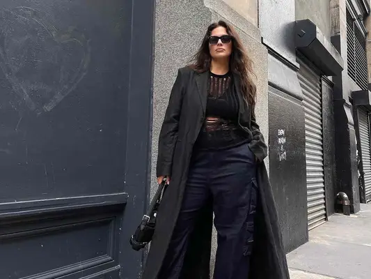 Ashley Graham wears an all-black outfit featuring open-knit sweater, cargos, longline trench coat, handbag, and sunglasses