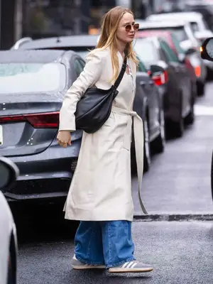 Jennifer Lawrence wearing a tan trench coat, black crossbody bag, and gray Adidas Sambas
