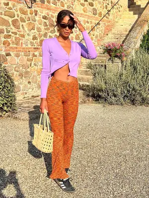 Laura Harrier wearing a bright purple cardigan, orange knit pants, and black Adidas Sambas