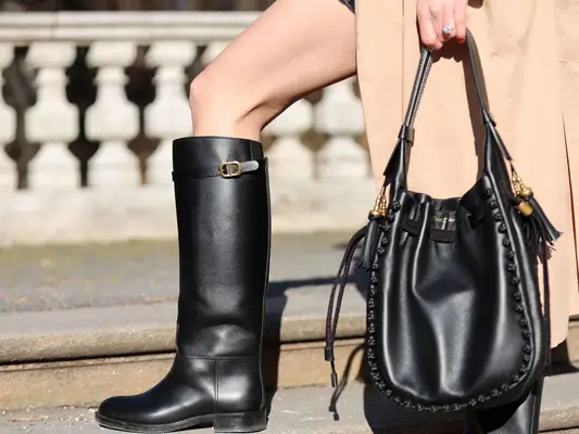Close-up of woman wearing Dior riding boots, oversized bag, and tan trench coat