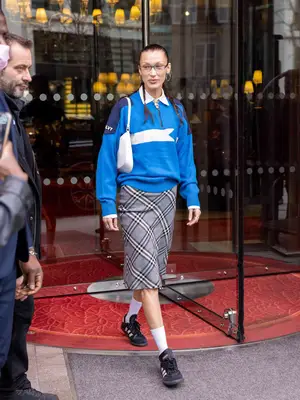 Bella Hadid wears a plaid skirt, collared quarter-zip, Adidas Samba sneakers, white socks, white shoulder bag, and glasses