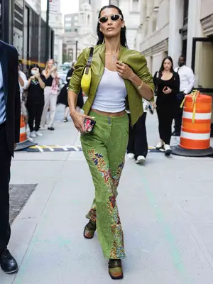Bella Hadid wears a white tank top, green embroidered trousers, a green leather jacket, oval sunglasses, and a yellow shoulder bag