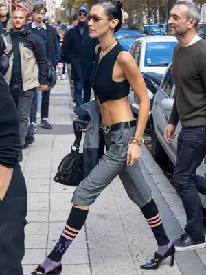 Bella Hadid wears a vest top, knee-length trousers, knee-high striped socks, loafer heels, and tiny sunglasses
