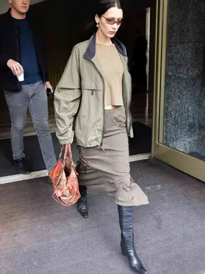 Bella Hadid wears a patterned top, khaki-colored midi skirt, all-weather jacket, leather boots, colorful bag, and sunglasses