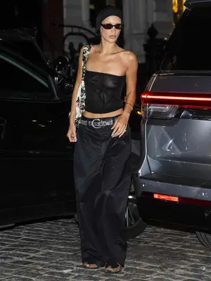 Bella Hadid wears a sheer black tube top, baggy pants, a patterned bag, tiny sunglasses, and a headband