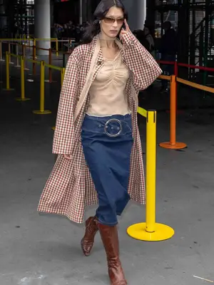 Bella Hadid wears a nude blouse, denim midi skirt, brown leather boots, gingham coat, and skinny scarf