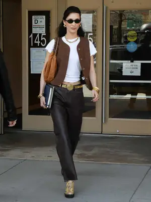 Bella Hadid wears a brown vest, white T-shirt, leather pants, loafers, and gold jewelry