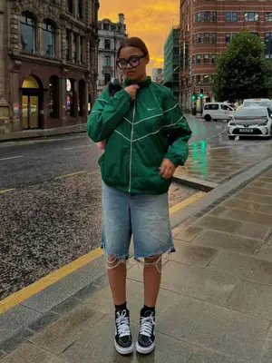 Madison Bailey wears a green Nike athletic bomber jacket, denim Bermuda shorts, black sneakers, and glasses