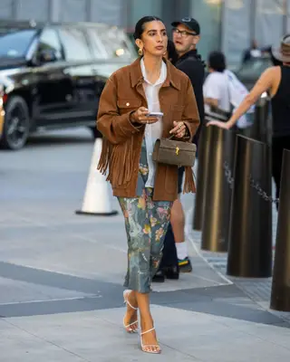 Woman wearing brown suede jacket with fringe, white button-down, floral midi skirt, and white strappy heels
