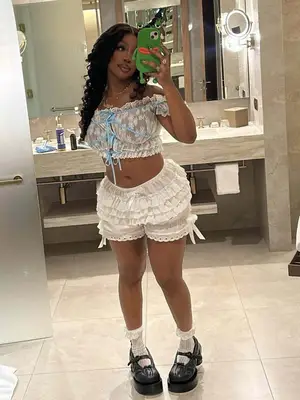 SZA is standing in front of a mirror wearing a white lace crop top, white bloomers, black mary jane heels, and lace frill socks