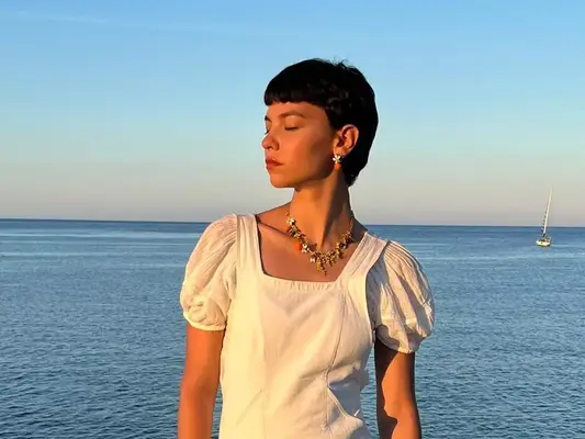 Woman wearing white puff-sleeve top and orange blossom-inspired jewelry by water