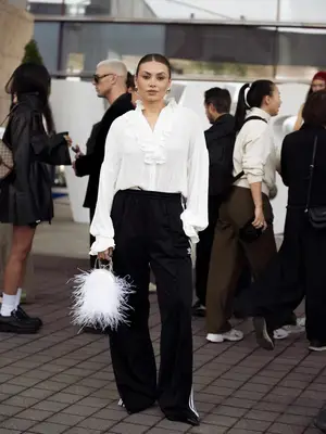 Person wears a ruffled white blouse with black track pants, pointed-toe heels and a feathered white bag