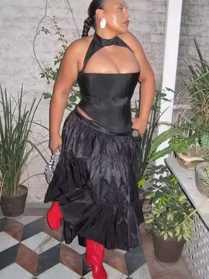 Paloma Elsesser wears a black corseted top and tiered maxi skirt with red boots, knit handbag, and oversized white earrings