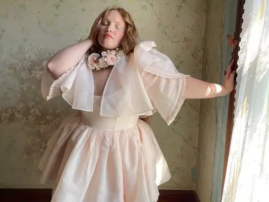 Model wearing light pink flouncy mini dress with capelet and rose details