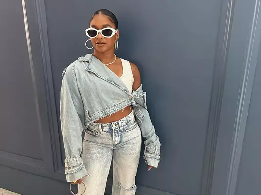 Jordan Chiles wears a white cropped tank top, frayed denim jacket, and light wash jeans with white sunglasses and silver hoops