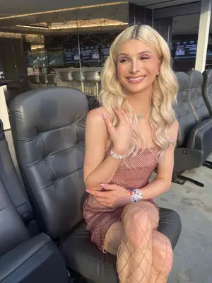 Dylan Mulvaney wears a sparkly pink dress, nude fishnet tights, sparkly hair and makeup, and beaded friendship bracelets