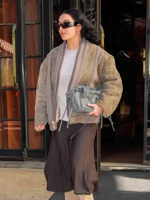Charli XCX carries an east-west handbag and wears sunglasses, an oversized sweater jacket, and wide leg pants